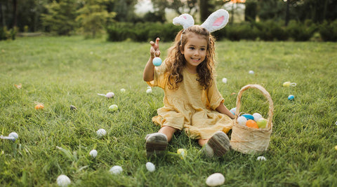 Menina com orelhas de coelho a segurar um cesto de ovos da Páscoa enquanto procura ovos na relva de um jardim verde. 