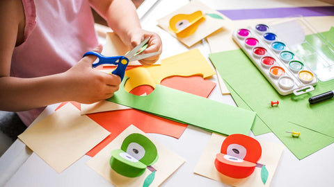 criança a recortar cartolinas coloridas com uma tesoura numa mesa com material de artes manuais